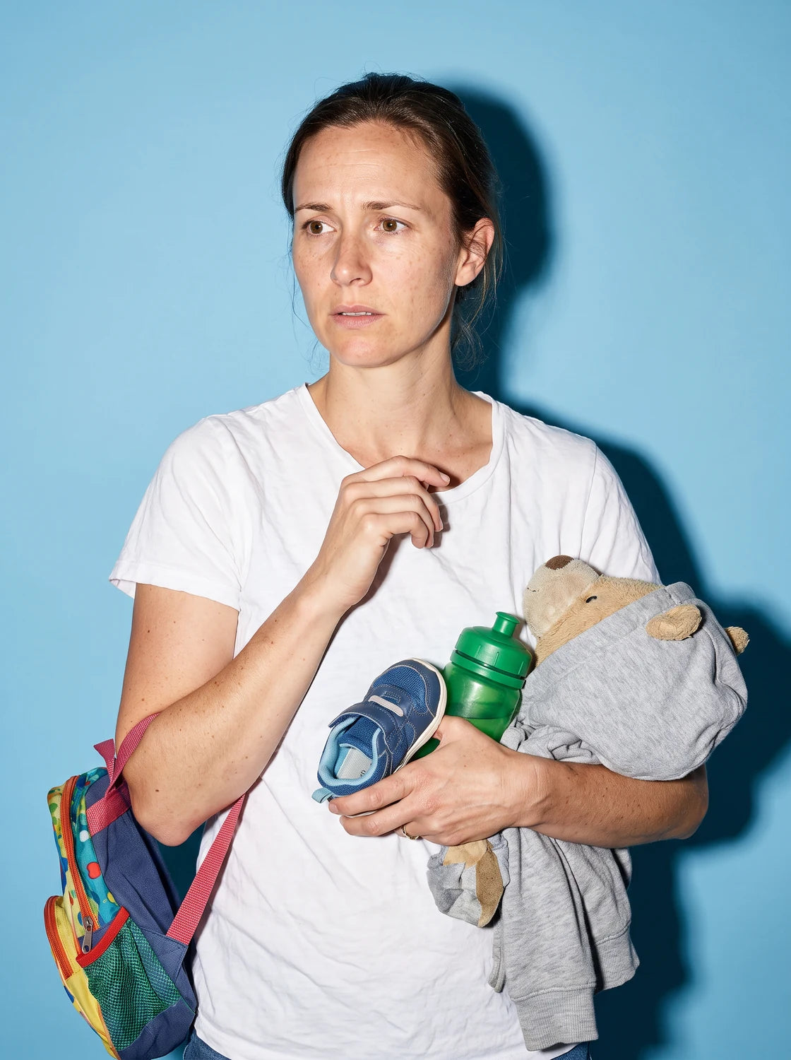 A mother carrying a kid's backpack, shoe, water bottle and stuffed bear, photographed with hard flash against a sky-blue backdrop
