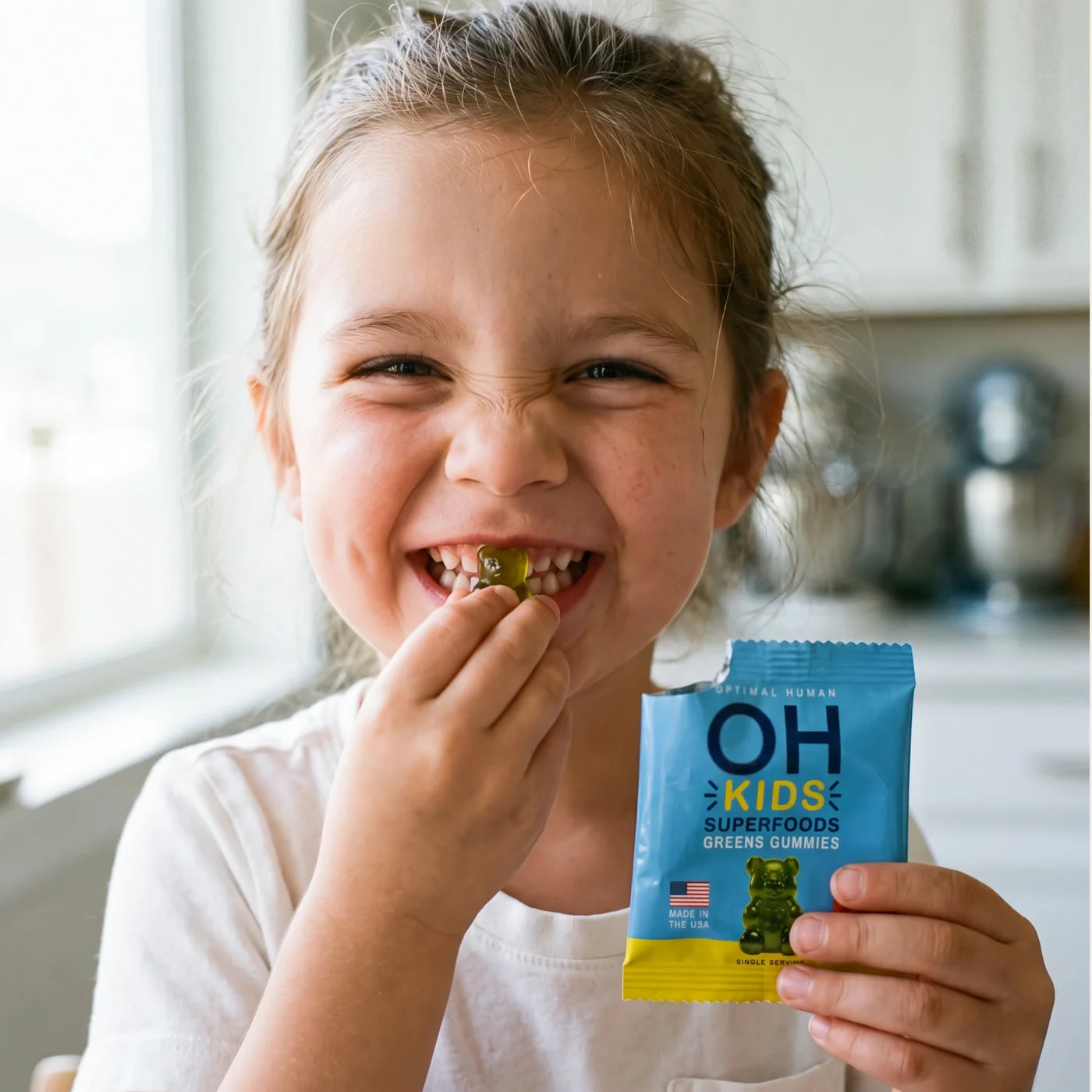 A young girl grinning with a green gummy in her teeth, holding a single blue OH Kids pack
