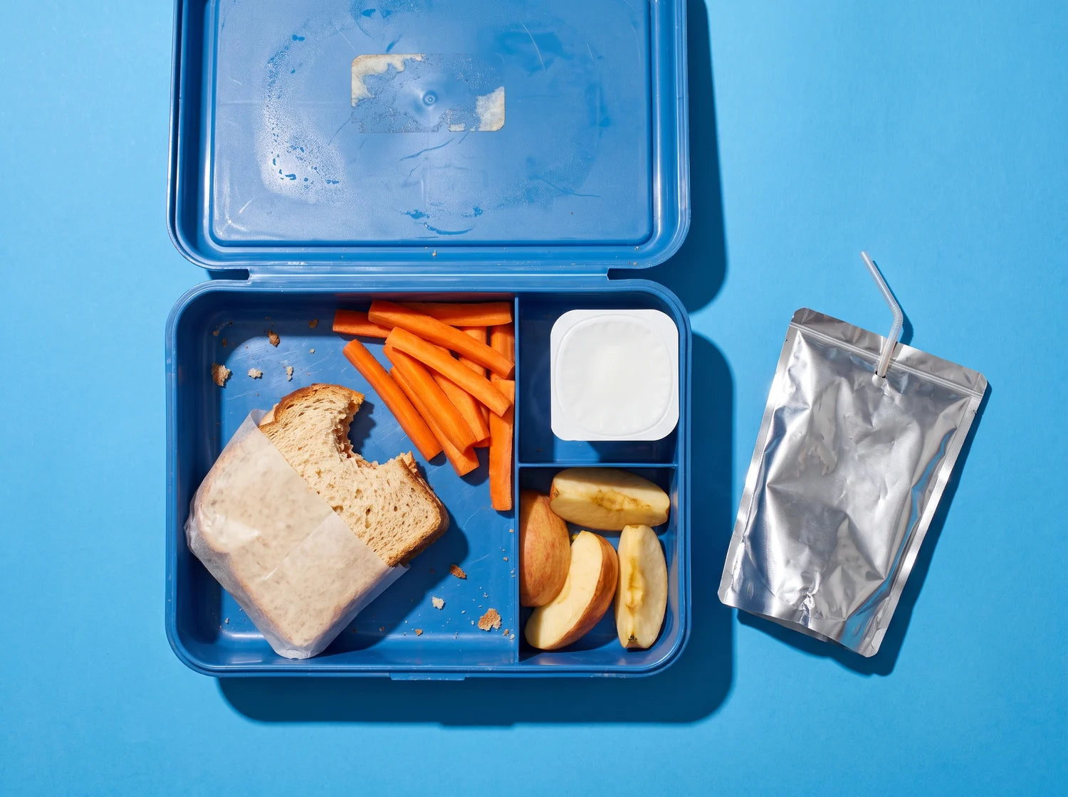 An opened school lunchbox come home with the sandwich barely bitten, carrots and apple slices untouched, and the drink pouch empty
