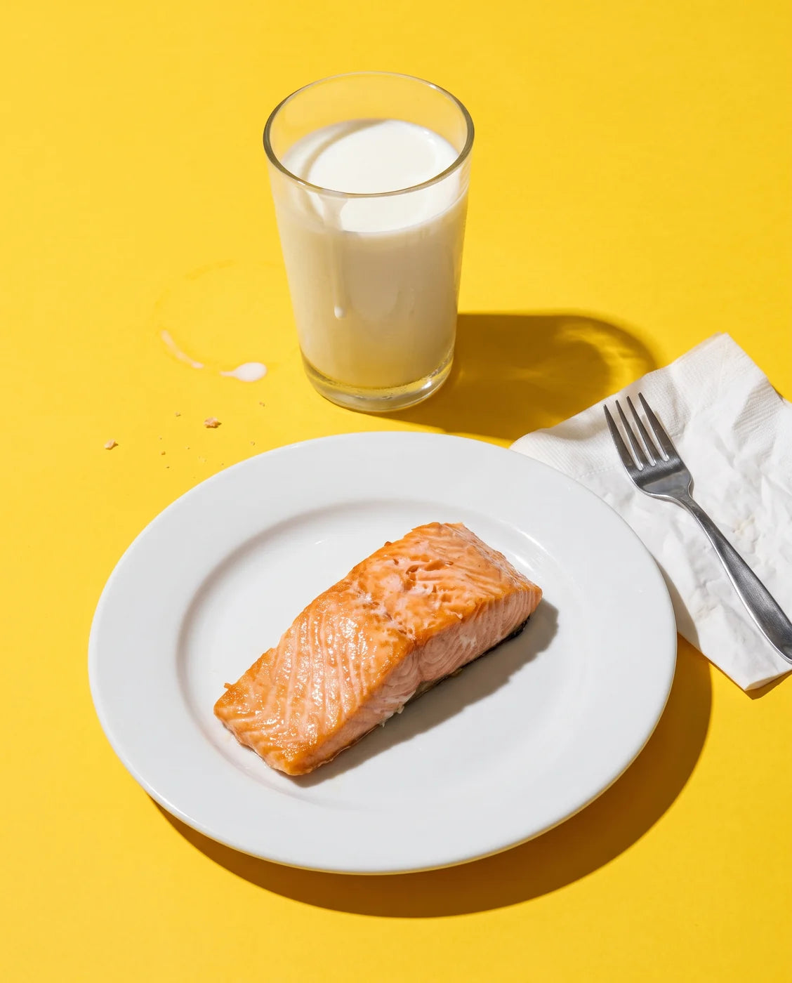 An untouched cooked salmon fillet on a plate beside a full glass of milk and an unused fork, shot with hard flash on a yellow backdrop