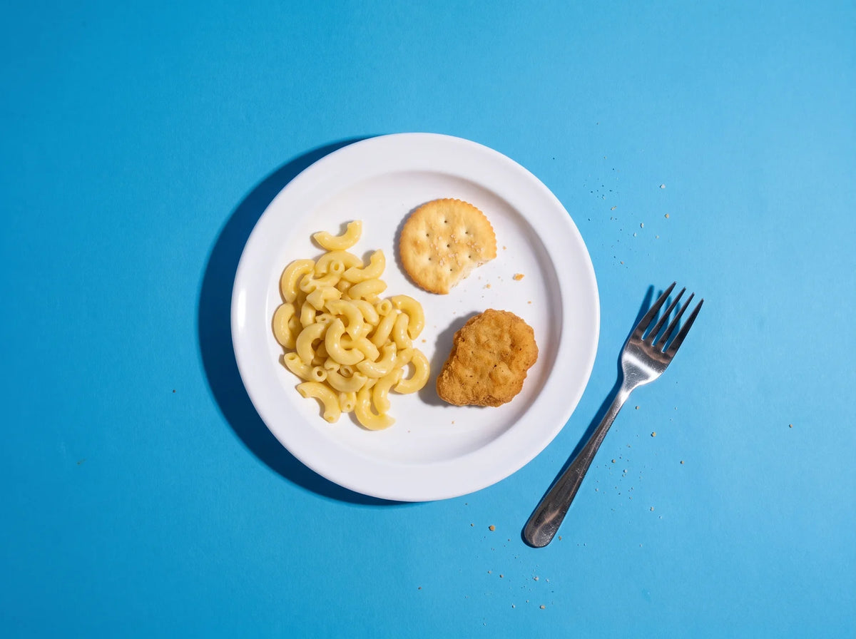 A kids dinner plate of plain pasta, a cracker and a chicken nugget