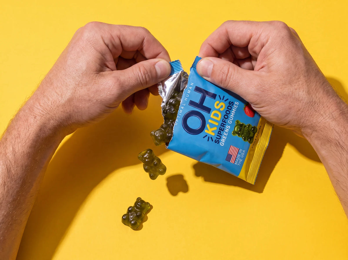 Hands tearing open a blue OH Kids Superfood Greens Gummies pack as green gummy bears spill out