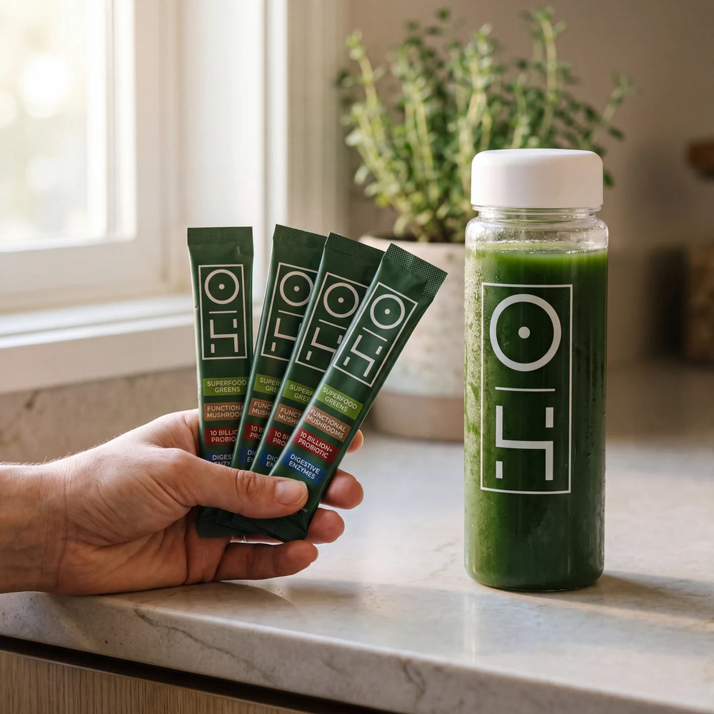 A hand holding four Optimal Human greens stick packs next to a bottle of greens on a kitchen counter