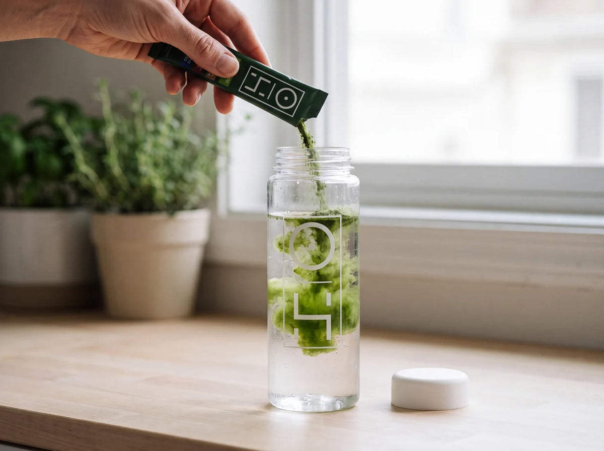 A hand pouring a Superfood Greens stick pack into a bottle of cold water