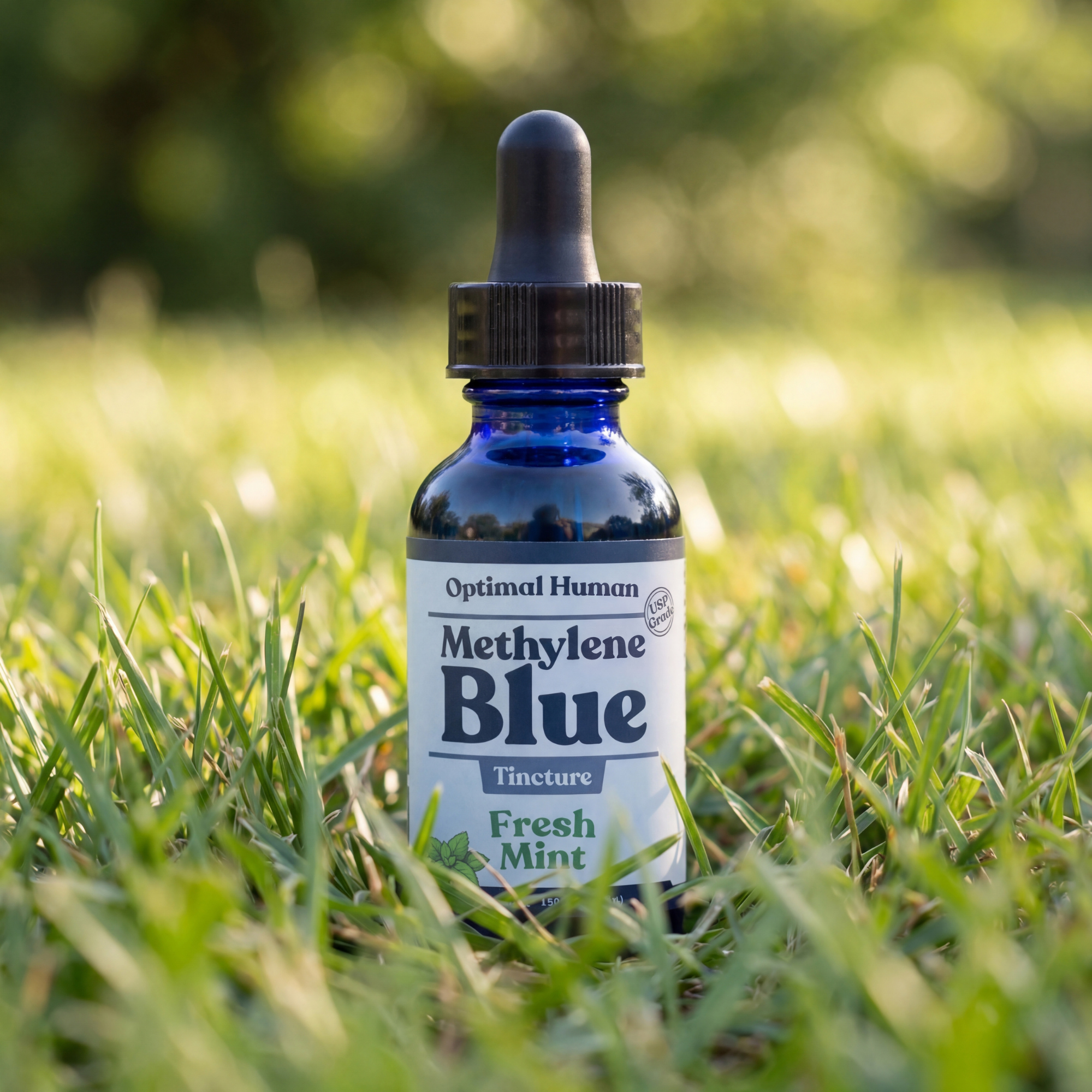 Optimal Human - USP Grade Methylene Blue (Fresh Mint) — Cobalt blue glass Optimal Human Methylene Blue Fresh Mint tincture bottle with black dropper cap standing in green grass at golden hour, soft bokeh nature background