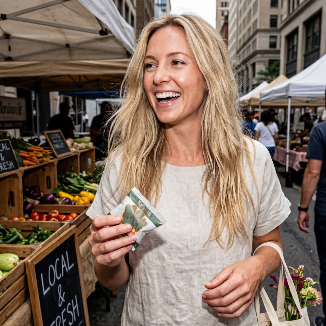 Optimal Human - Superfood Greens Gummies (30 Day Supply) — Lifestyle photo of blonde woman at outdoor farmers market holding a small Superfood Greens Gummies sachet, fresh produce crates and 'Local & Fresh' chalkboard signs in background