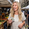 Optimal Human - Superfood Greens Gummies (30 Day Supply) — Lifestyle photo of blonde woman at outdoor farmers market holding a small Superfood Greens Gummies sachet, fresh produce crates and 'Local & Fresh' chalkboard signs in background