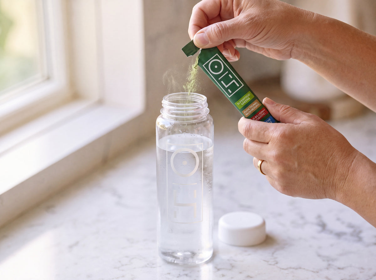 Hands tearing open an Optimal Human Superfood Greens stick over a bottle of cold water