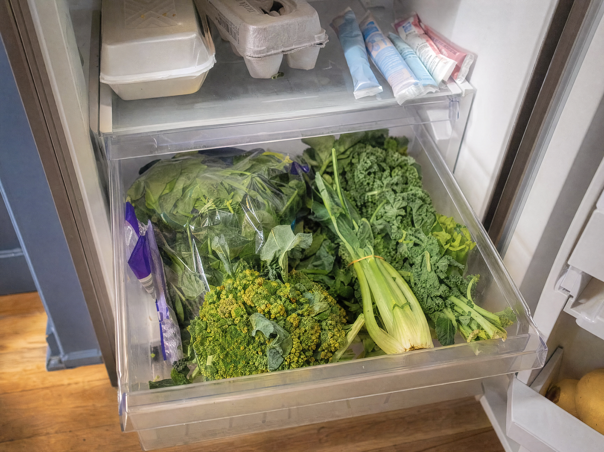 An open fridge crisper drawer full of spinach, broccoli and kale going soft