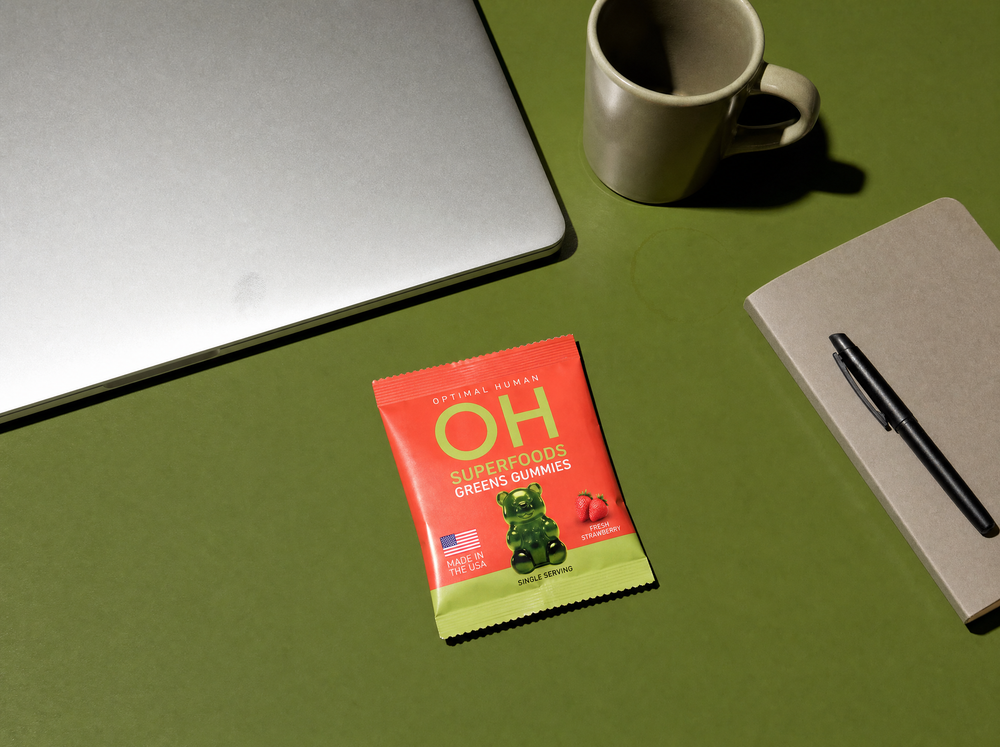 A single-serve pack of greens gummies on a desk beside a laptop and a mug