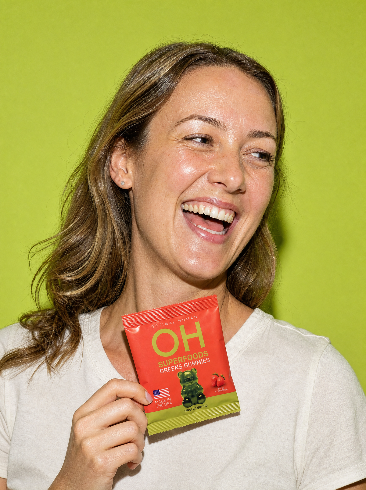 A woman laughing while holding a single-serve pack of greens gummies