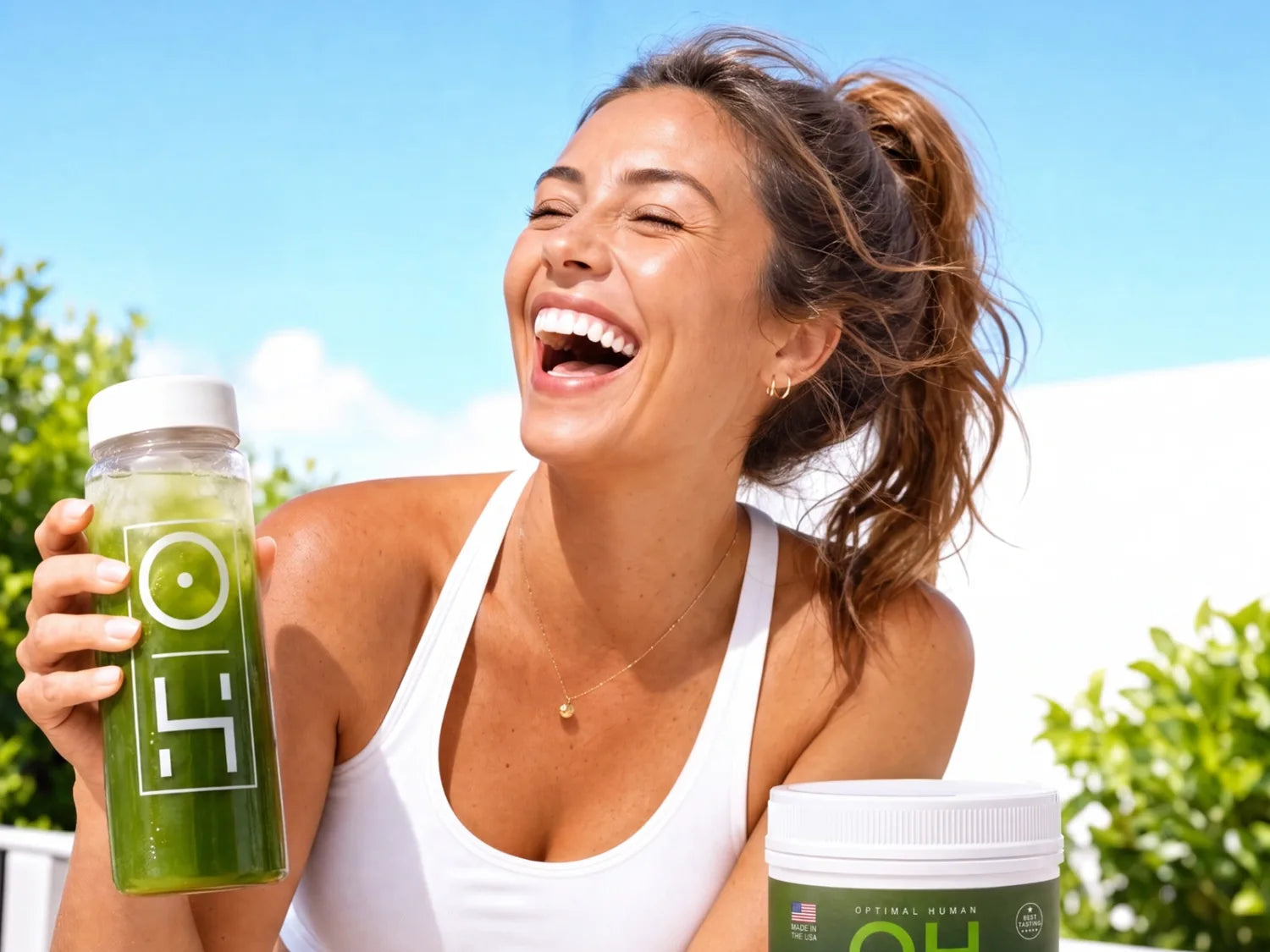 A woman laughing outdoors in the sun, holding a clear OH bottle of the mixed greens drink, with the tub beside her