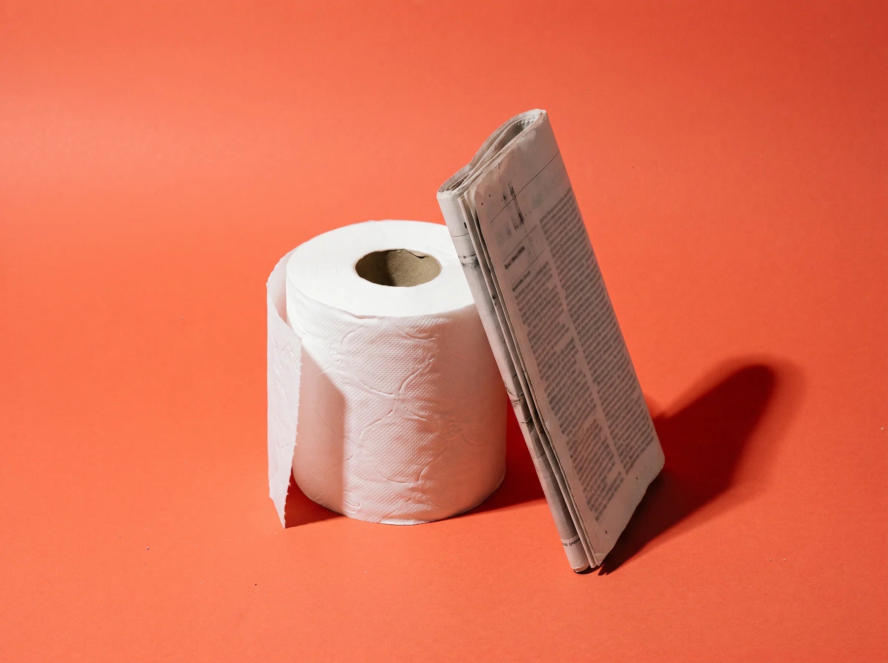 A roll of plain white toilet paper standing on end with a folded newspaper leaning against it, studio still life on coral paper