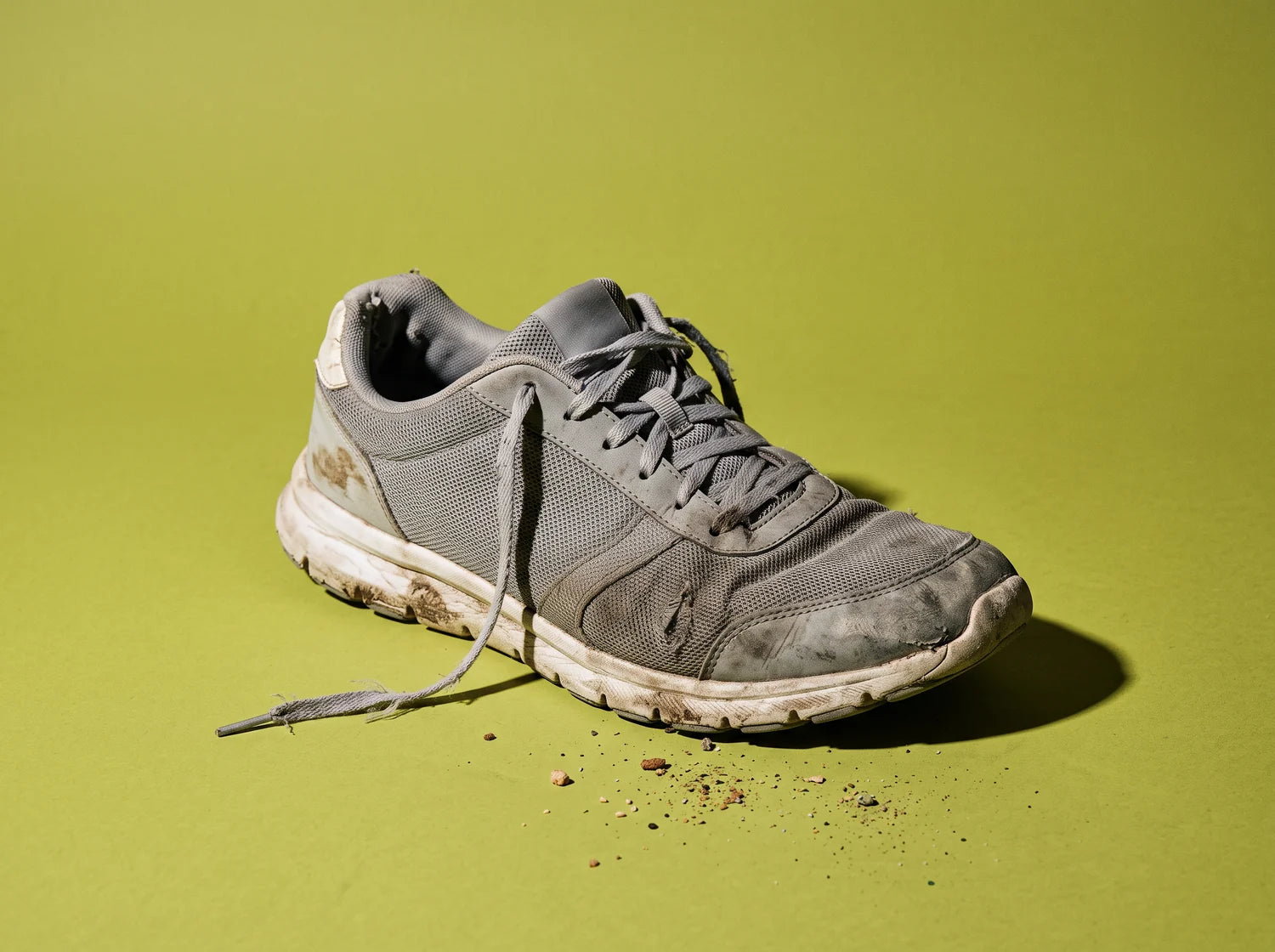 One well-worn grey walking shoe with a loosened lace and dirt in the tread, studio still life on lime paper