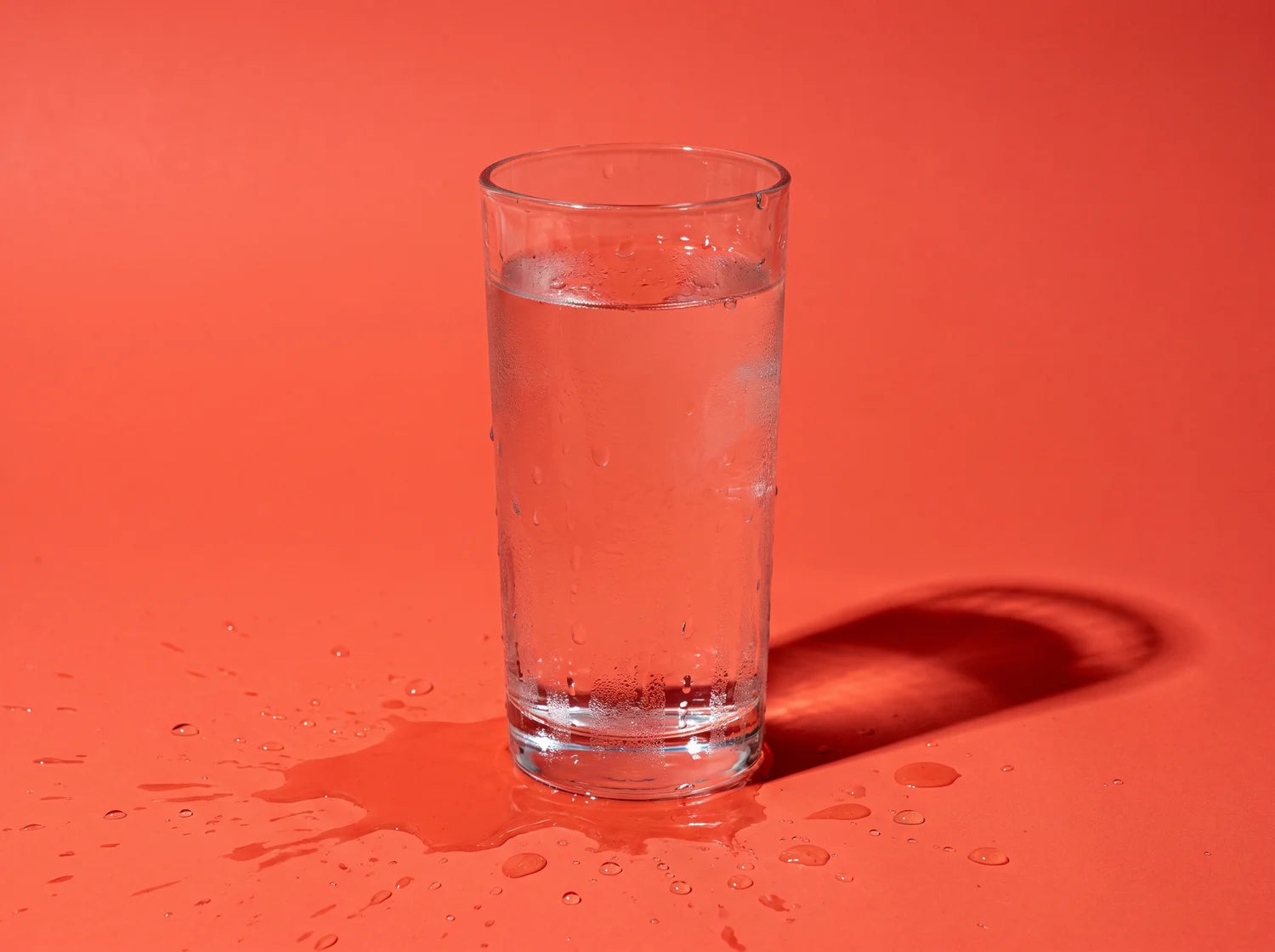 A tall glass of cold water beaded with condensation, water pooling on the paper around it, studio still life on coral paper