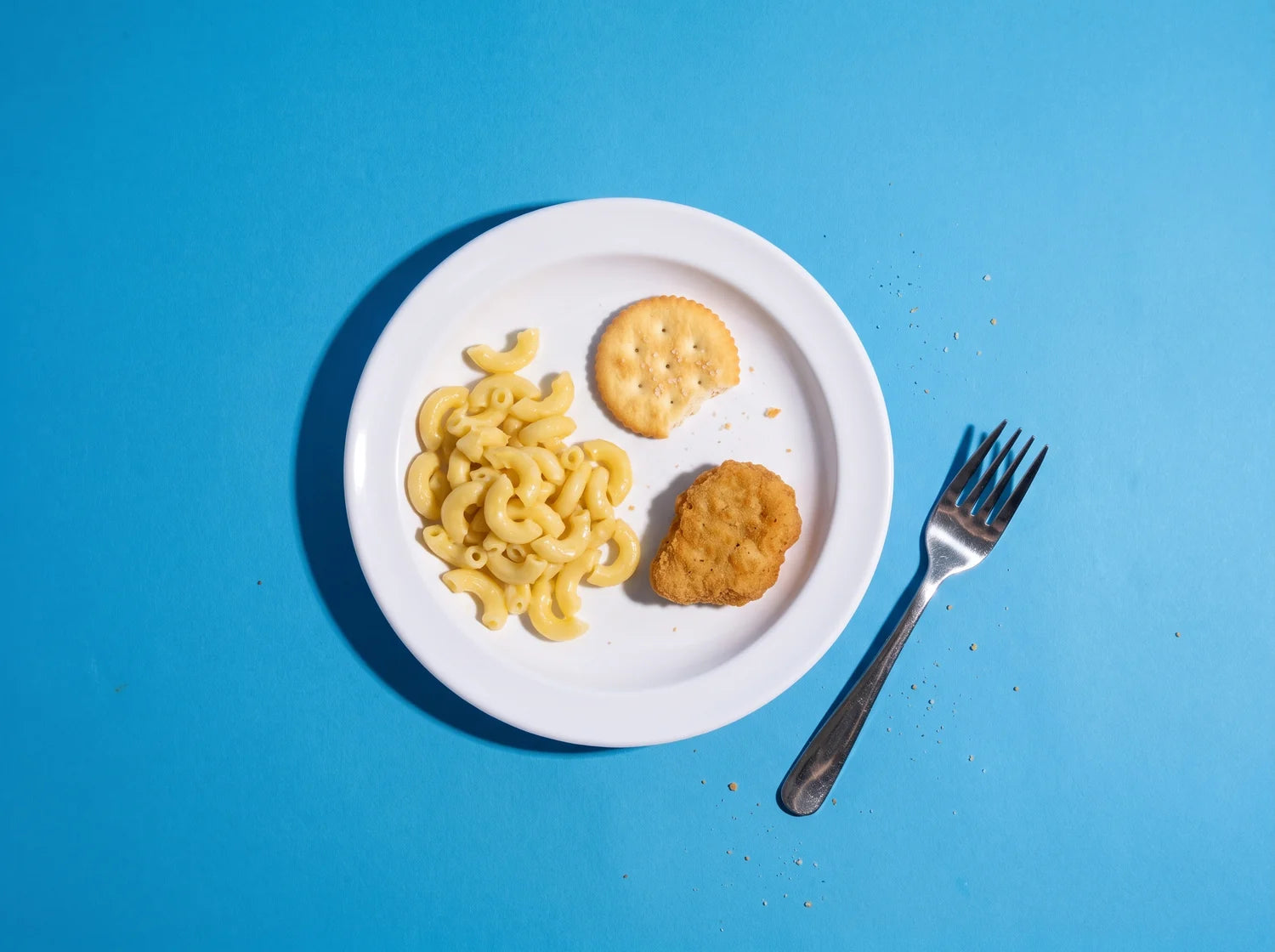 A child's plate holding only plain pasta, one cracker and a chicken nugget, shot with hard flash on a blue backdrop