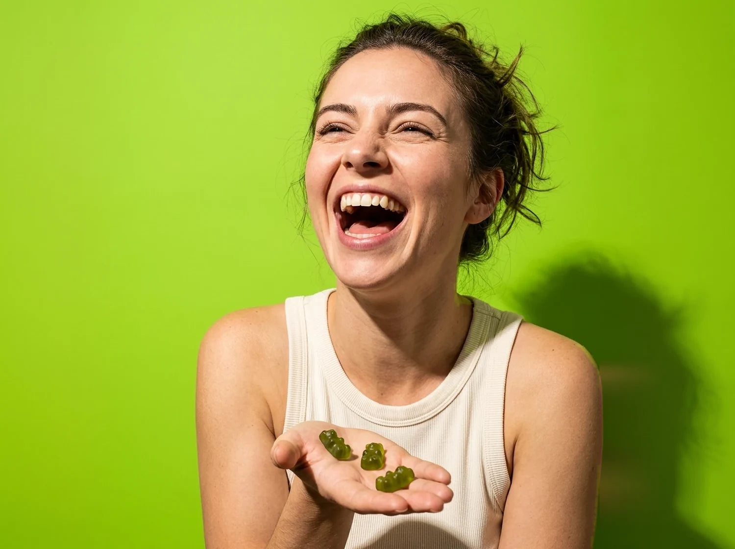 A woman laughing with a handful of green Optimal Human gummy bears in her open palm, on lime paper