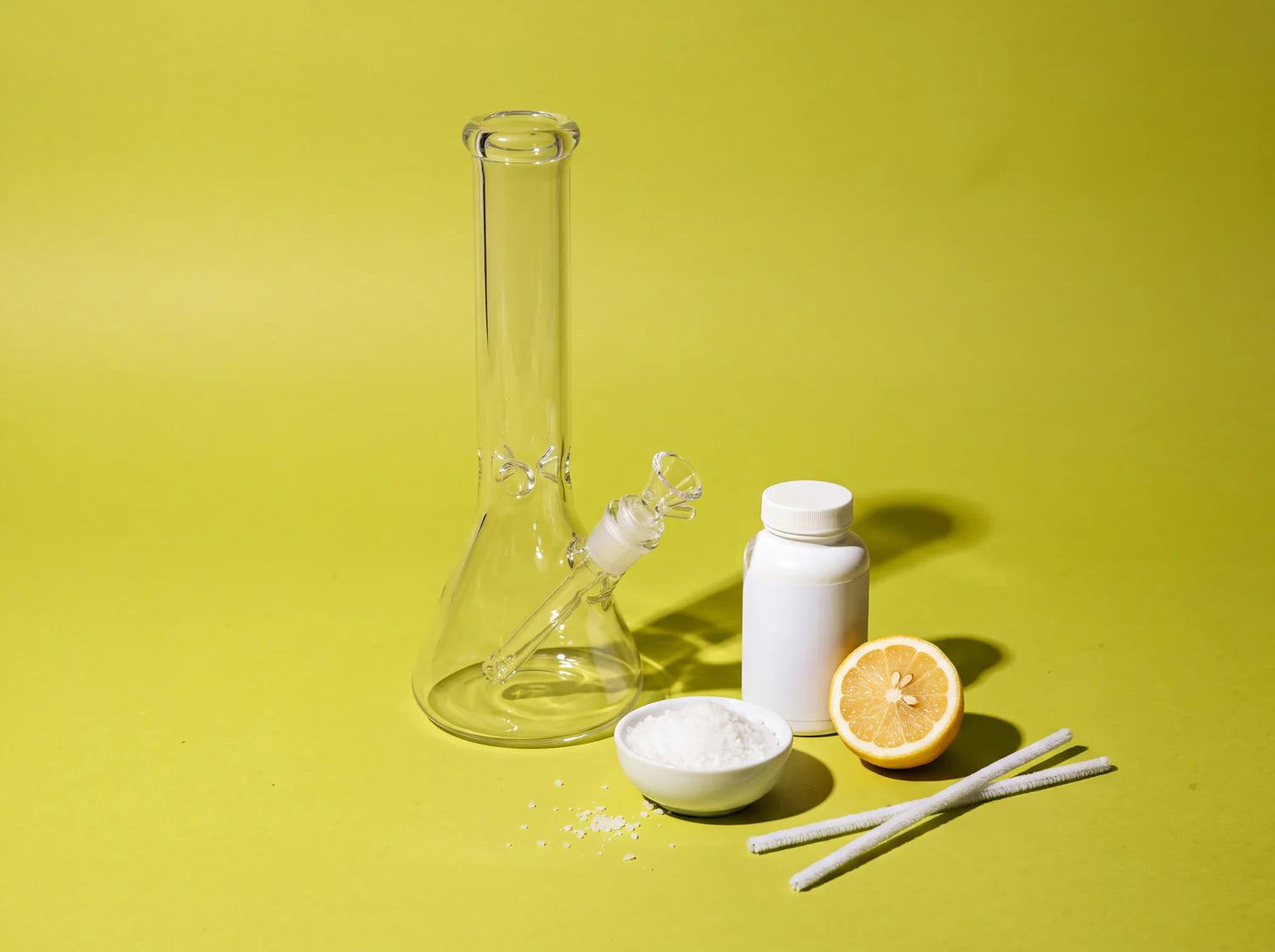 A spotless clear glass water pipe beside a bowl of coarse salt, a plain bottle, half a lemon and pipe cleaners, on lime paper