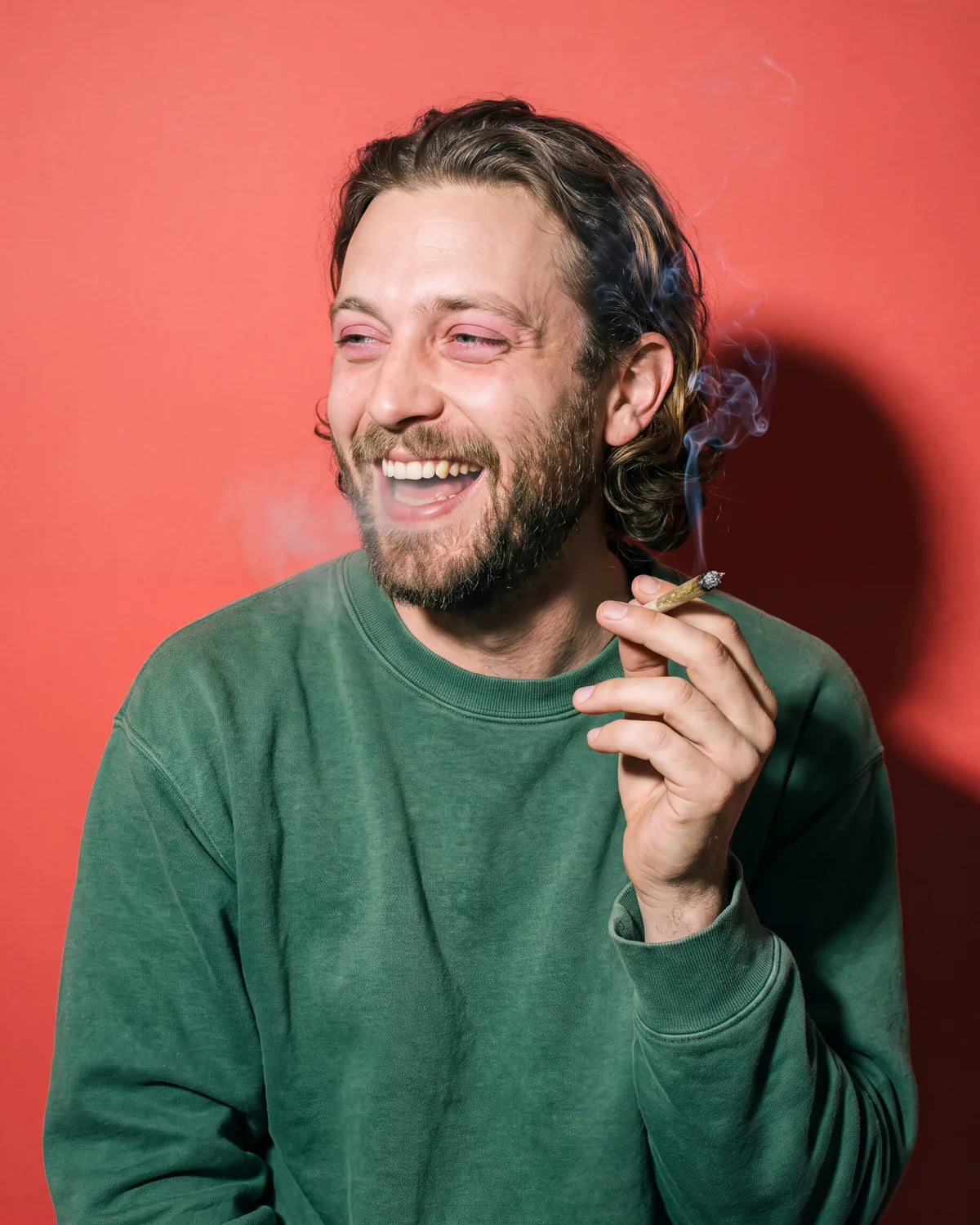 A bearded man in a green sweatshirt laughing with a friend just out of frame, on coral paper