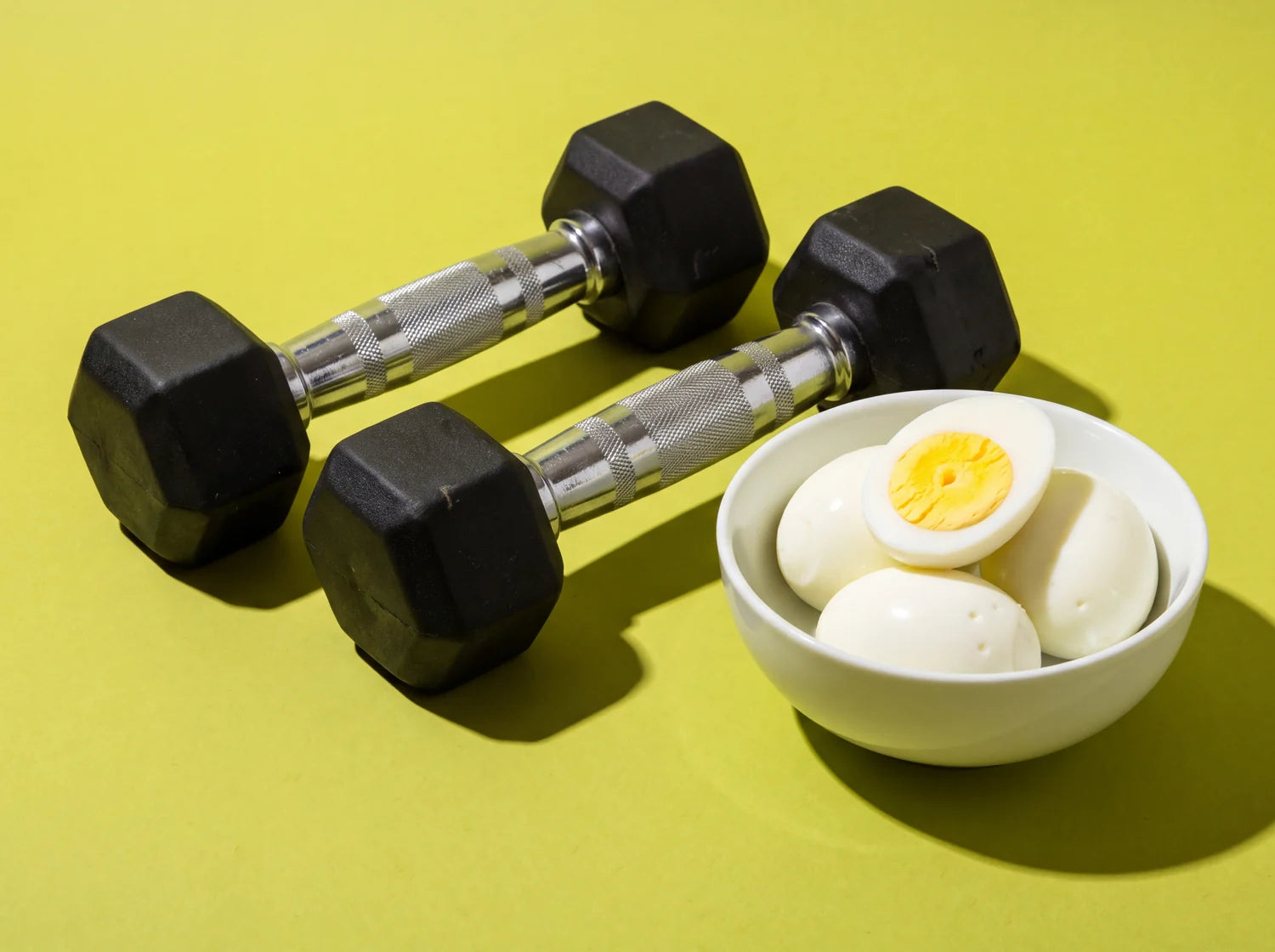 A pair of black dumbbells beside a bowl of hard-boiled eggs, studio still life on lime paper