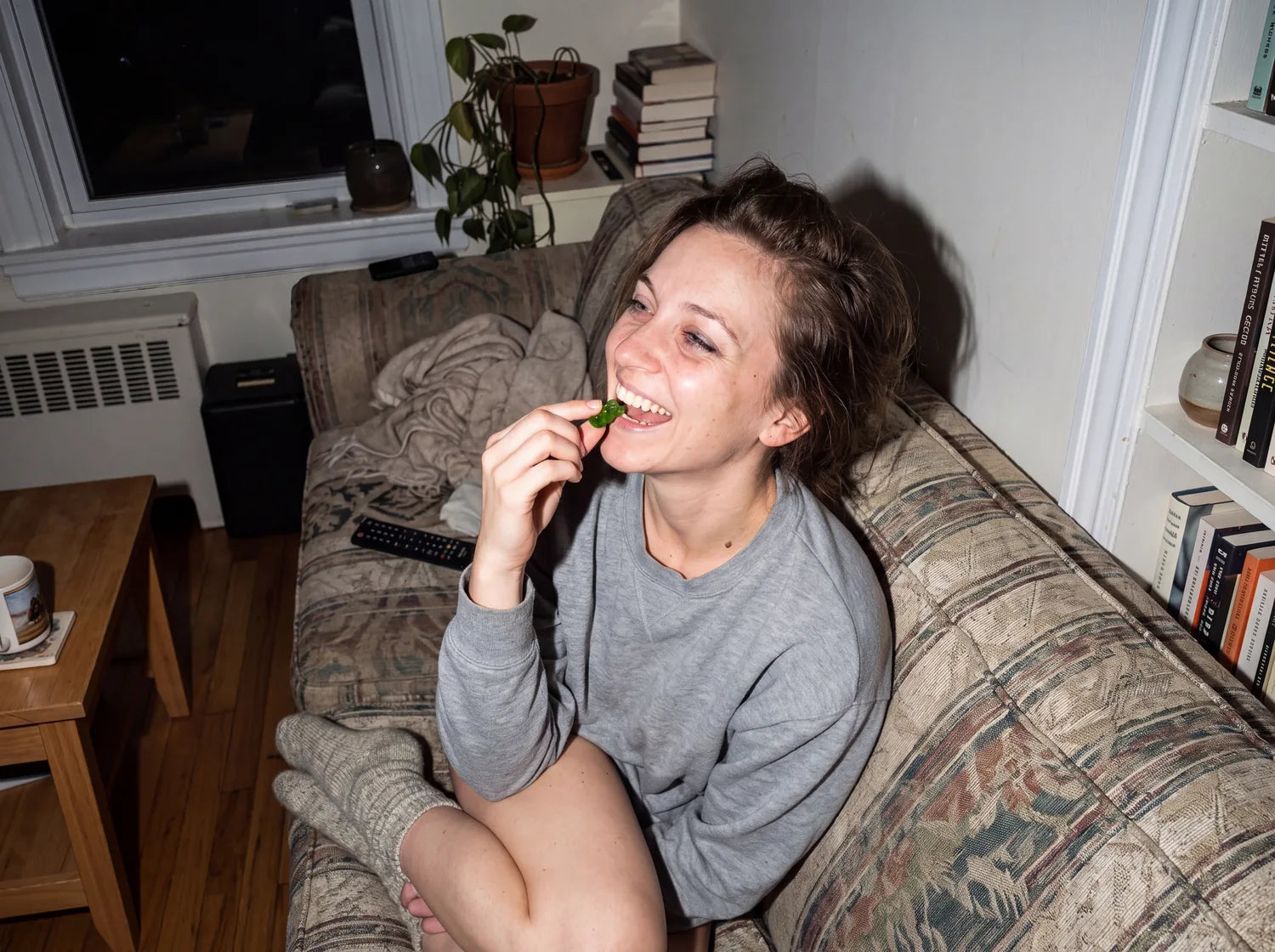 A woman laughing on a worn couch at night while she bites a green Optimal Human gummy bear