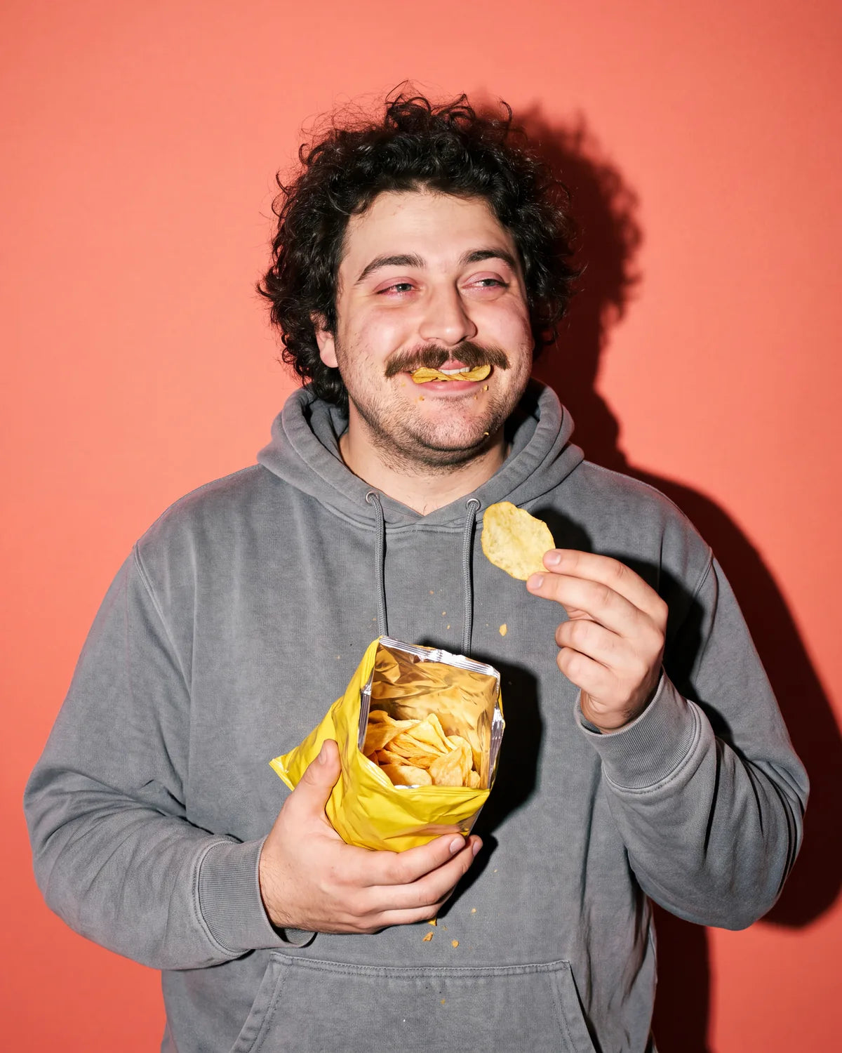 A very happy, very stoned man with a potato chip between his lips, holding an open bag of chips, on coral paper