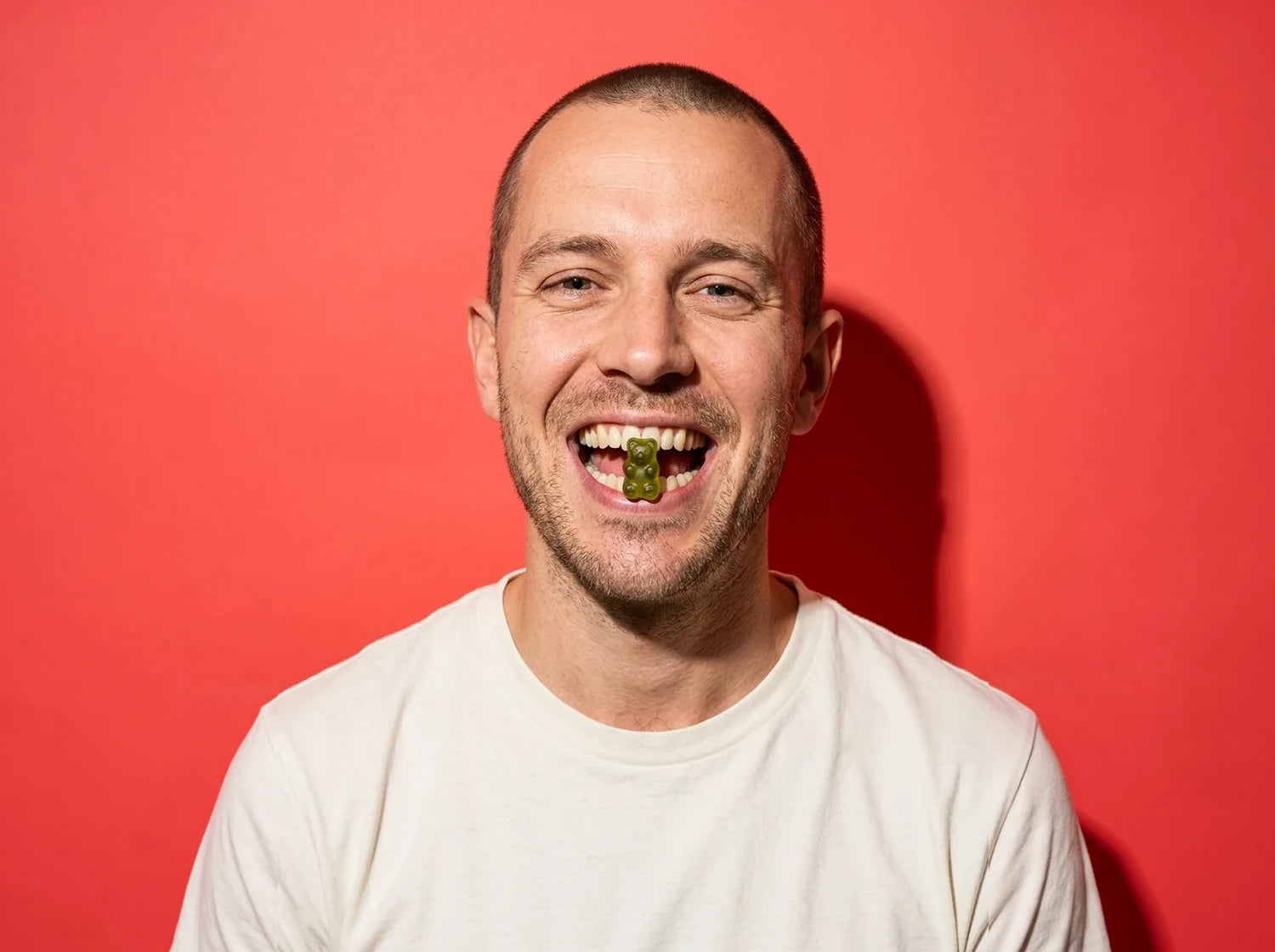 A smiling man holding a green Optimal Human gummy bear between his teeth, on coral paper