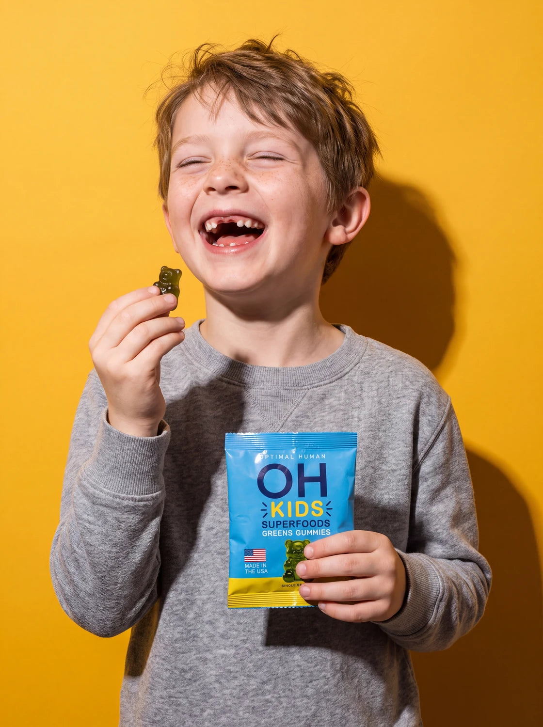 A freckled boy laughing with a green gummy in one hand and a blue OH Kids pack in the other