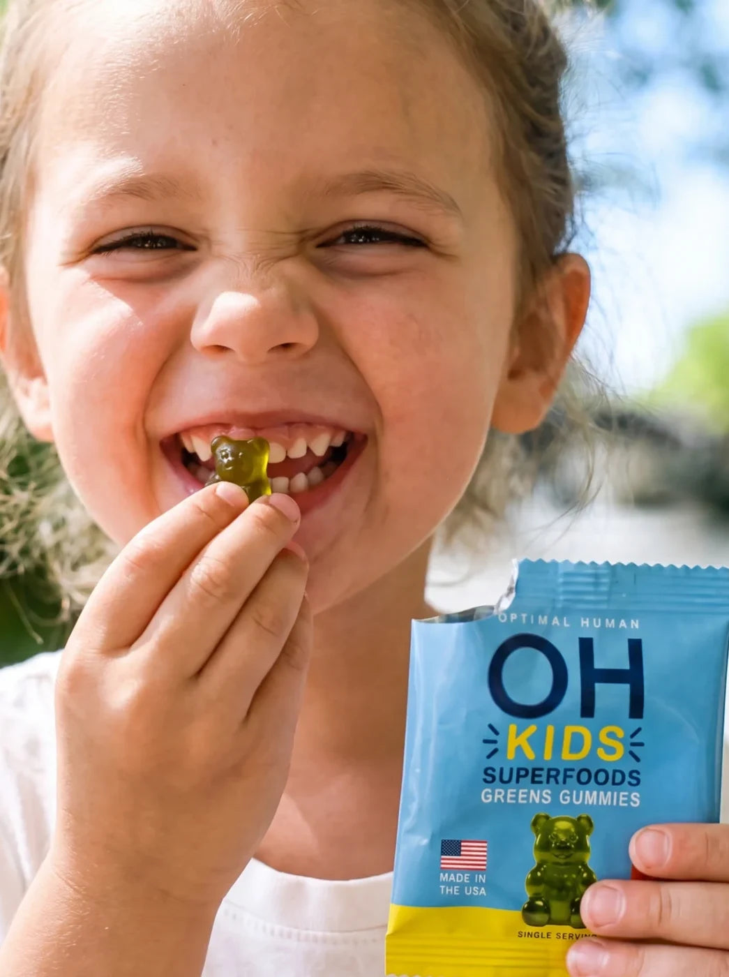 A little girl grinning with a green gummy in her teeth, holding an OH Kids pack