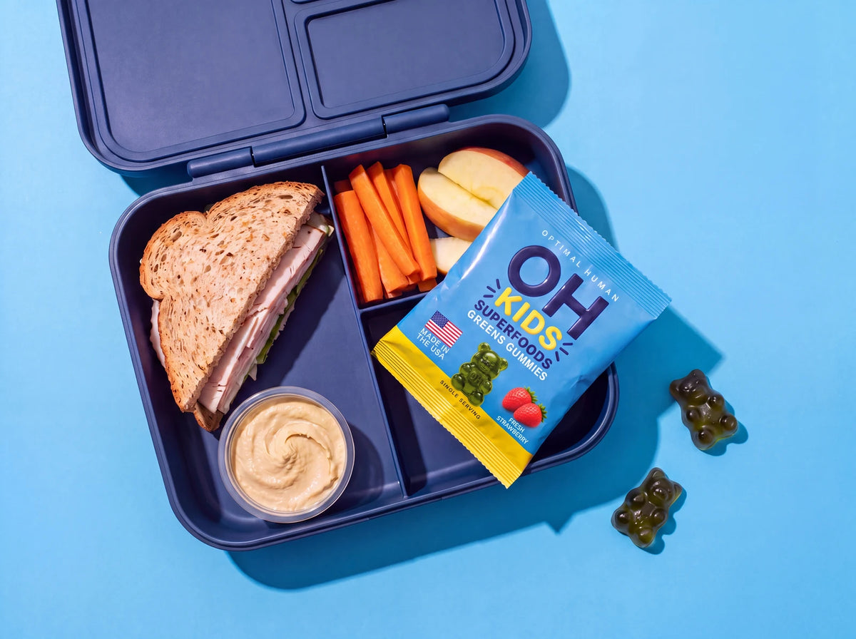 A packed school lunchbox with a turkey sandwich, carrots, apple slices, hummus and a blue OH Kids gummies pack