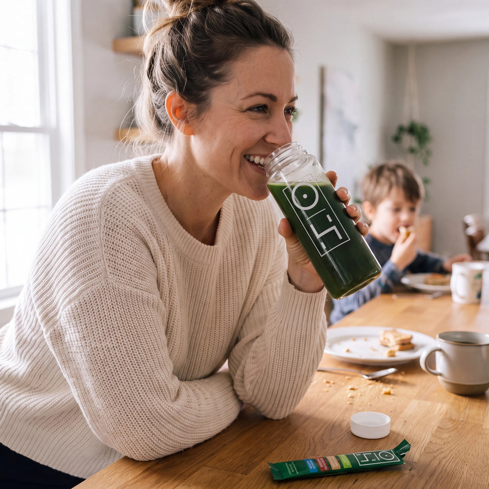 A mom laughing as she sips Superfood Greens from the bottle in her kitchen, her son behind her