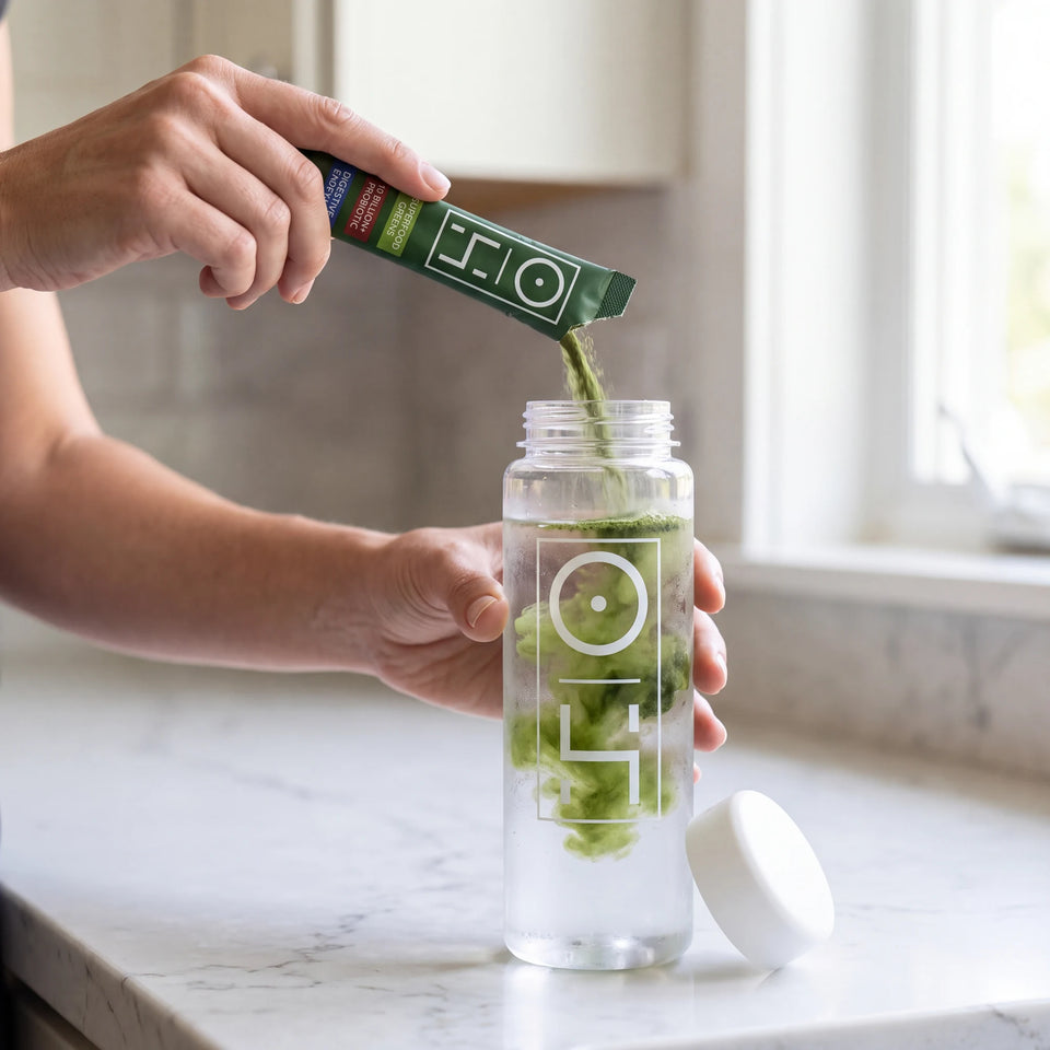 A hand pouring a Superfood Greens stick pack into a bottle of cold water