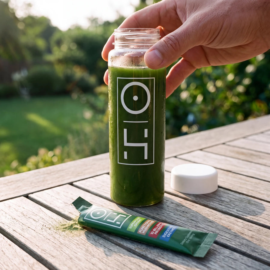 A hand lifting a bottle of bright green Superfood Greens on a sunny garden table, a stick pack beside it