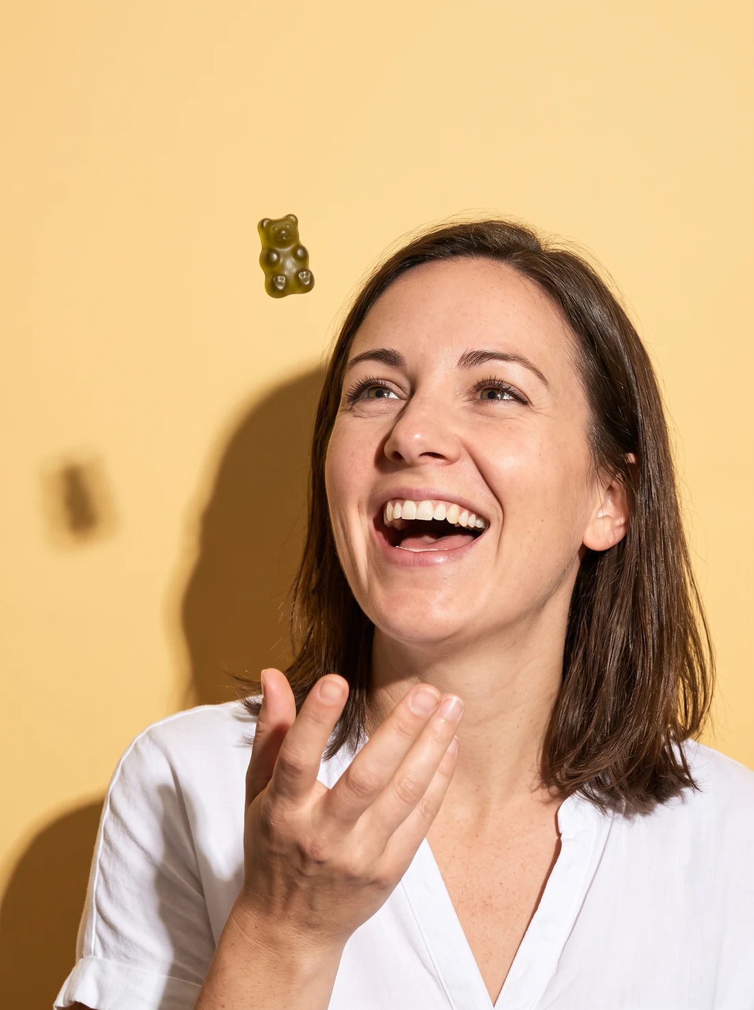 A smiling woman tossing a green gummy toward her mouth